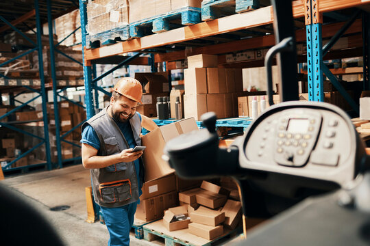Smiling warehouse worker using smartphone while carrying box in storage aisle