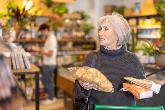 Store visitor, senior woman in sales area choosing pasta. Client make choice of assortment, select, buy noodle product. Customer read label, examine customer information, view package content