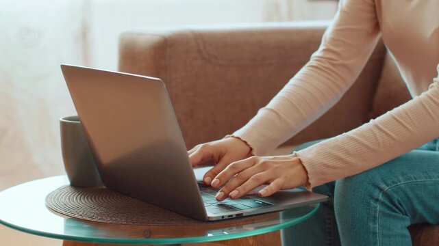 A woman with light skin is sitting on a couch, typing on a laptop. She has a cup of coffee on a round table beside her. Sunlight enters the room, creating a warm atmosphere.
