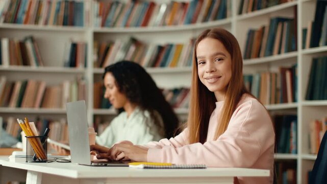 Two young women are focused on their laptops in a library setting. One woman with long red hair types, while another woman with dark curly hair is engaged in her work.