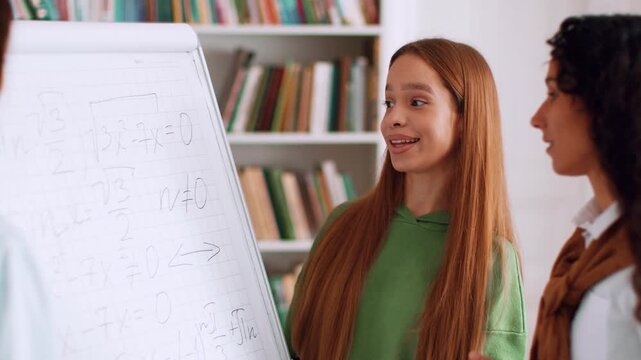 Three students collaborate in a classroom environment. One student explains a math concept using a whiteboard while the others listen attentively. Bookshelves are visible in the background.