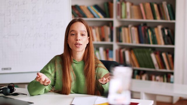 A young woman with long brown hair is speaking while seated at a desk. She is teaching in a study space with shelves of books in the background and writing on a whiteboard.