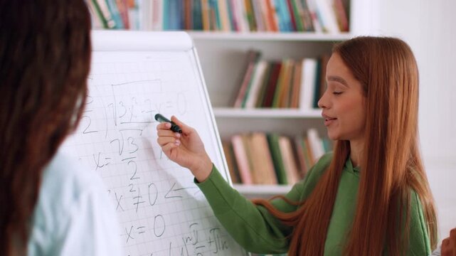 A young girl with long red hair writes on a whiteboard while teaching a lesson in a classroom. Shelves filled with books are visible in the background. She is engaged and smiling while explaining.