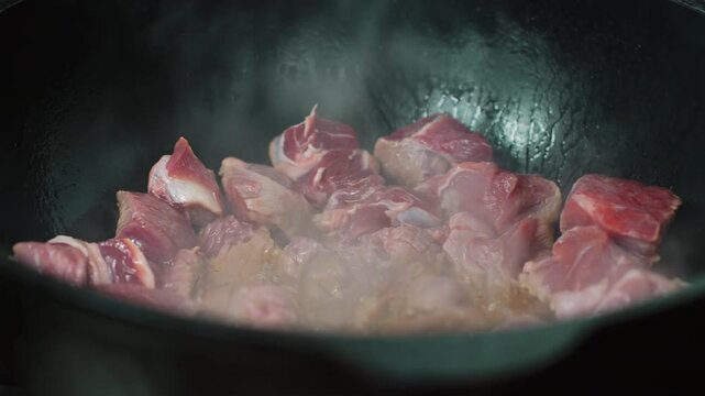 Multiple raw beef chunk pieces arranged at the bottom of a large black cast iron wok with oil, beginning of braising or stewing process, dark background