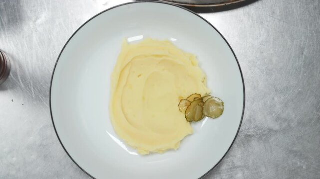 Chef plates creamy mashed potato on a white plate, places two golden fried meat cutlet on top and adds pickle slice as garnish, top view on stainless steel kitchen surface