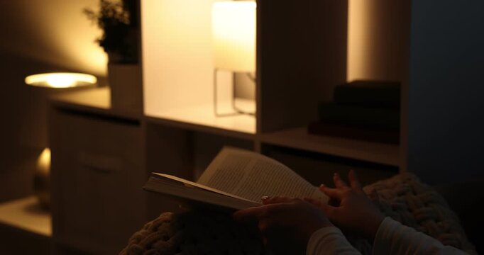 Woman flipping through book pages with illuminated night lamps at home, closeup