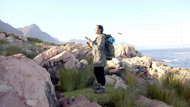 Vertical video: Facing hiker in parka with pack and mat on cliff checking phone VIBES appearing