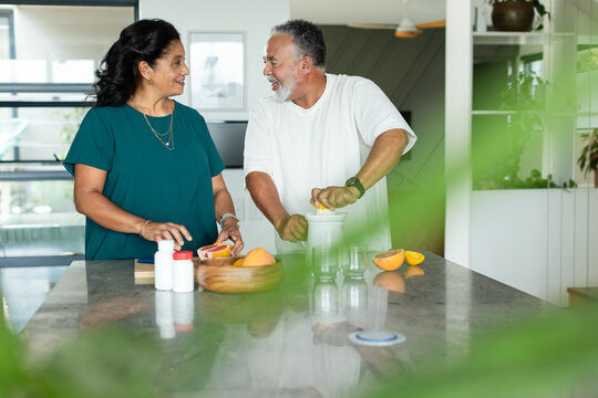 Senior couple standing at stone kitchen island making juice with citrus press, woman in teal blouse