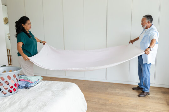 African American couple holding large pale sheet while folding laundry near bed and wardrobe