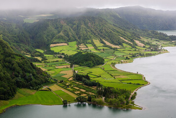 Volcanic crater lakes surrounded by lush greenery. Twin lagoons in Sete Cidades, Azores. Rolling hills and pastures under cloudy sky. Rural landscape with scattered villages in Portugal. © Eugene Ga