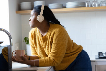 African American woman leaning at sink wearing mustard knit and white headset holding peach mug © wavebreak3