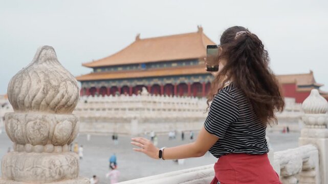 Caucasian vlogger filming forbidden city selfie with smartphone, animated head turn, ornate red pavilion backdrop, busy plaza activity, bracelet on wrist, candid capture for social content