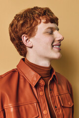 Young man with red hair wearing a brown leather jacket and turtleneck smiling and looking sideways on beige background, casual style and profile portrait for fashion concept. © SHOTPRIME STUDIO