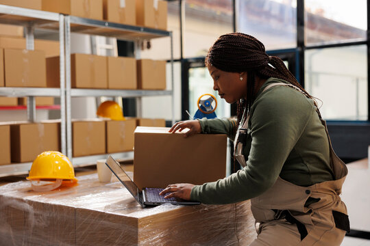African american worker checking customers orders on laptop, preparing packages using cardboard boxes in storage room. Warehouse manager wearing industrial overall during inventory