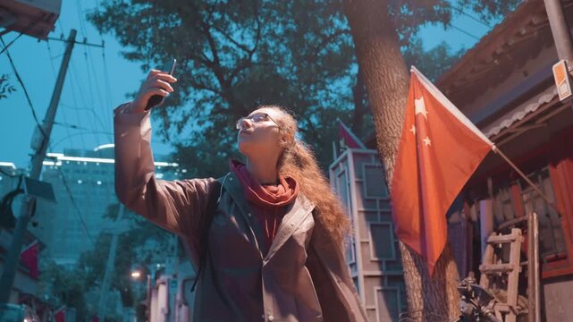 Woman walking narrow alley at dusk, navigating lanternlit lane with smartphone in hand, wearing coat and scarf, fluttering flags and soft streetlamp glow, cinematic tealorange palette, reflective mood
