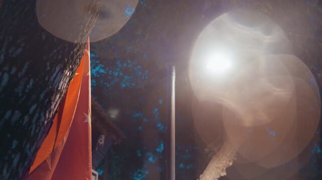 Night lamp red flag closeup fluttering over eaves, intense lens flare and circular bokeh, warm glow and light haze, fabric texture visible against quiet urban roofline, evokes nostalgia and solitary