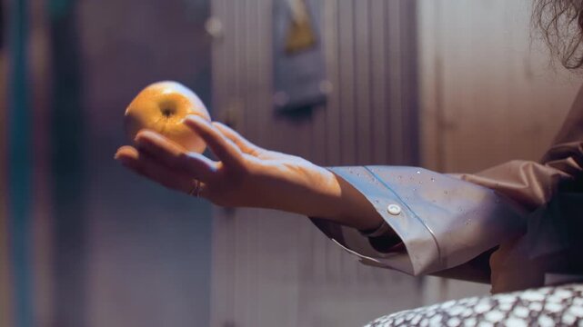 Hand releasing orange in dim light, levitating motion over palm, industrial backdrop, neon highlights, sleeve detail, slow float, shadowed performer energy, suspenseful urban atmosphere