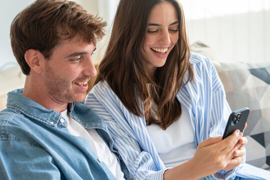 A young couple sits on a couch smiling and looking at a smartphone together, sharing a happy, relaxed moment at home while browsing photos or messages in a bright living room.