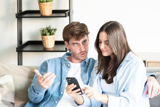 Young couple sits closely on a sofa at home, sharing a smartphone and discussing plans with relaxed, intimate expressions that convey connection, collaboration and modern lifestyle.