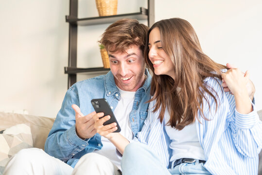 A young couple sits close on a sofa, smiling and surprised as they look at a smartphone together, sharing a joyful moment at home that conveys connection, affection, and modern communication.