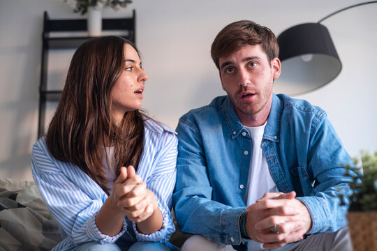 Young couple sitting together on a sofa at home, engaged in a tense conversation. Expressions show frustration, concern and distance as they discuss relationship issues and try to communicate.
