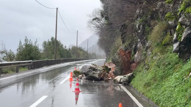 Driving around rockfall on wet mountain road in rain, landslide hazard, traffic danger. Traffic cones and warning sign indicate a landslide hazard as the vehicle carefully drives around the obstacle. 
