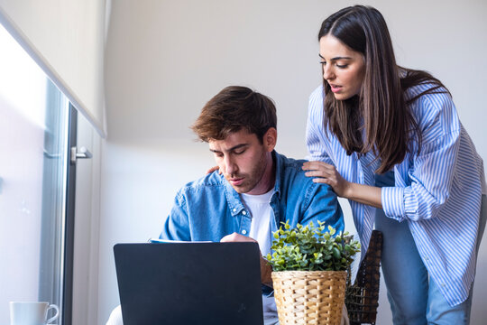Young couple sit at a laptop in a bright home office, reviewing finances and paperwork. The woman leans in to comfort and support her partner during a stressful moment of decision making.