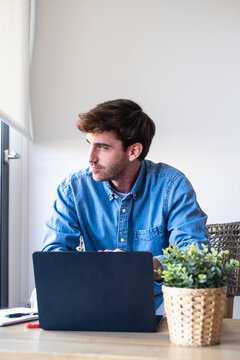 Young man sits at a home desk with a laptop and plant, working remotely and looking thoughtfully out the window. Casual, focused mood conveying remote work, productivity and modern home office life.