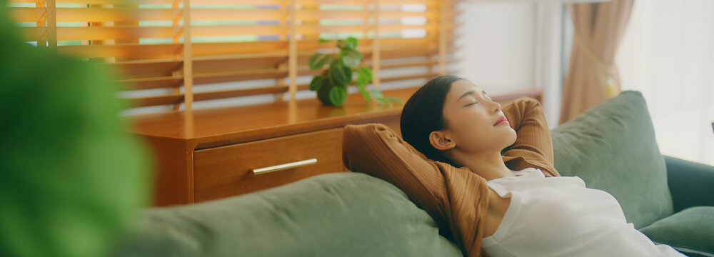 Relaxed young Asian woman enjoying rest on comfortable sofa, calm attractive girl relaxing on couch, breathing fresh air with eyes closed, meditating at home