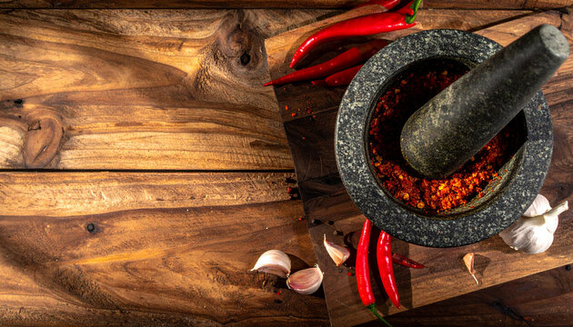 Top-down view of a stone mortar and pestle filled with crushed red chili surrounded by fresh peppers and garlic cloves on a dark wooden rustic surface with dramatic shadows