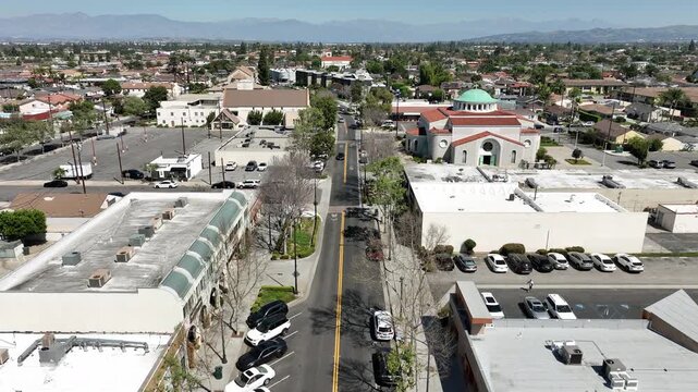 Aerial drone view of Downtown Downey, California, featuring tourists walking between sports bars, restaurants, and shopping centers. Bright daytime lighting highlights pedestrian activity