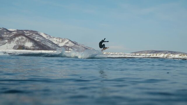 Surfer riding wave with snowy mountains along cold ocean coastline, rearview cut back getting air, slow motion