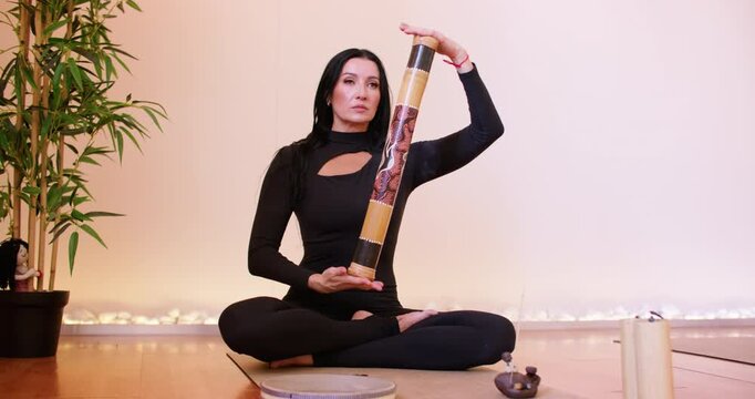 Yoga practice. Sound healing. Woman sitting in lotus position holding bamboo rainstick painted with ethnic pattern in meditation process.