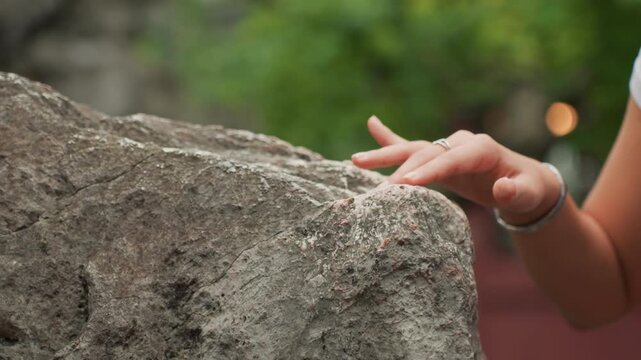Closeup hand touching rough rock, gentle fingers trace granulated surface with bracelet and ring visible, shallow depth of field and soft green bokeh background, sensory study of weathered mineral