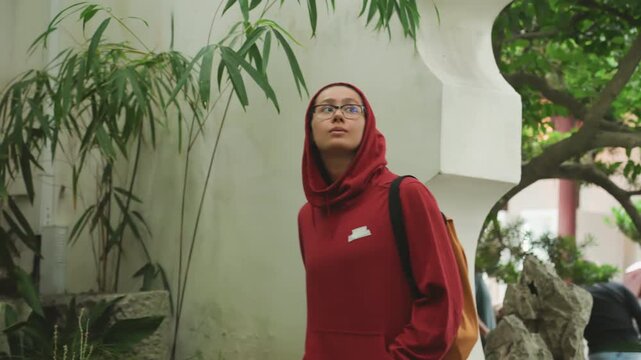 Asian woman red hoodie exploring classical garden, student strolling past curved white wall and mossy rocks, backpack slung over shoulder, glasses catching dappled sunlight, bamboo and pavilion