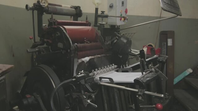 Authentic photo of an old letterpress printing machine in a workshop environment. Mechanical parts and paper feed system visible. Traditional analog printing process. Real industrial scene, not AI gen