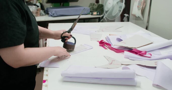 Dressmaking process. Tailor desk. Woman hand cutting curved garment textile pattern with heavy weight scissors in studio interior.
