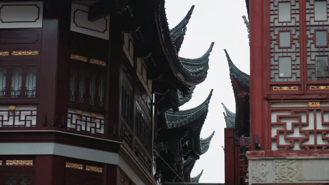 Ornate red chinese temple roof detail showing layered upturned eaves, carved lattice windows, dark lacquer beams, decorative brackets beneath overcast sky narrow courtyard and historic facade create