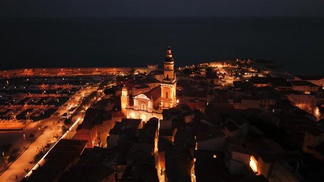 Menton Basilique Saint-Michel Archange at night, bell tower over orange rooftops, dark sea, France, drone