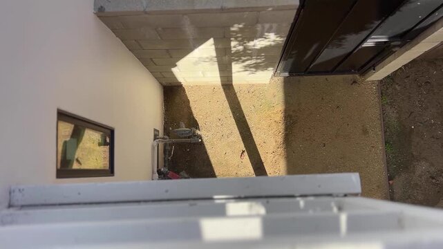 White apartment balcony in Brentwood during a sunny morning, with sun rays hitting the wall near a black door. Static top-down view.