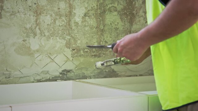 A tradesperson in high-visibility clothing uses a hammer and chisel to remove old plaster from a wall above a cabinet frame during a renovation.
