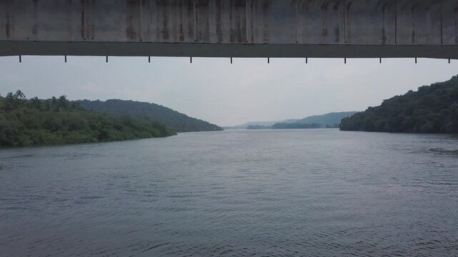 Concrete bridge spanning the Karli River with forested hills and backwaters in Devbag, Malvan, Maharashtra, India. The drone moves beneath the bridge structure towards the calm river channel.