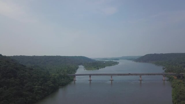 Concrete bridge spanning calm backwaters of Karli River near Devbag and Malvan in Maharashtra, India. Green forested hills line both riverbanks under overcast sky.