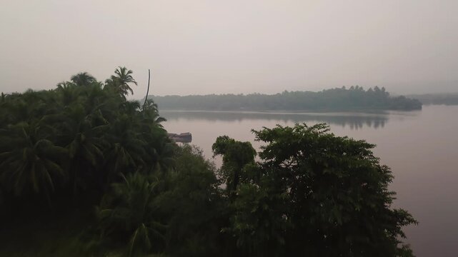 Camera rises over farm fields to reveal fishing boats on the tranquil Gad River backwaters in Malvan, Maharashtra, India during early morning fog and mist.