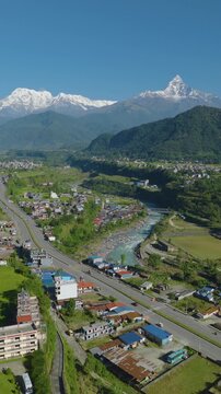 Vertical drone shot of a colorful village in Pokhara Nepal beside Prithvi Highway near forest and the Gandaki River with Machhapuchhre and Annapurna peaks rising.