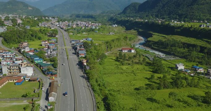 Aerial reveal shot of Prithvi Highway passing a village and green forest beside the Gandaki River with Annapurna and Machhapuchhre scenic mountain range rising in Pokhara Nepal.