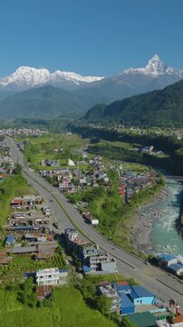 Aerial 9:16 portrait drone shot of rural Pokhara with Prithvi Highway running beside the blue Gandaki River through green fields and forest with Annapurna and Machhapuchhre peaks rising in Nepal.