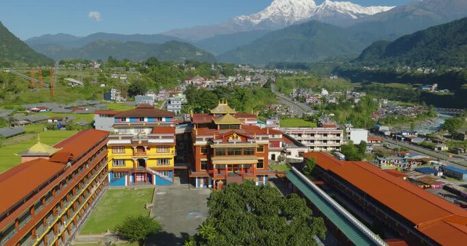 Aerial shot of a Buddhist monastery surrounded by village, green fields, forest and the Gandaki River with Annapurna and Machhapuchhre peaks rising in the background in Pokhara Nepal.