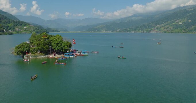 Aerial shot of Tal Barahi Temple n Phewa Lake Nepal with colorful boats floating on blue water and Nepal flag visible under a cloudy sky in Pokhara.