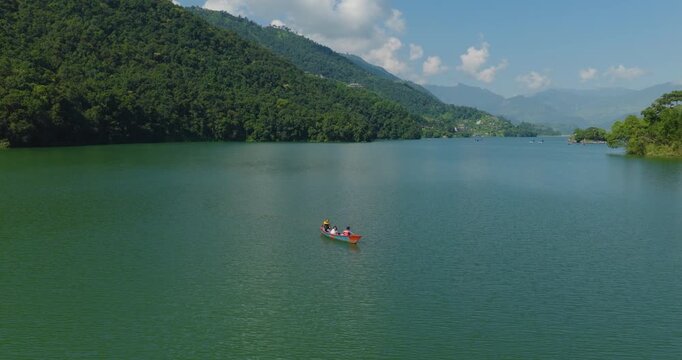 Aerial shot of three tourists rowing a boat on calm Phewa Lake Nepal Pokhara surrounded by green forest beneath a cloudy blue sky.